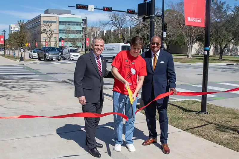 Harris County Commissioner Rodney Ellis and Michael Johnson, the University of Houston president’s chief of staff, on Tuesday, Feb. 25 hosted a ribbon-cutting ceremony to celebrate completion of the Cullen Boulevard project that runs through the heart of the Third Ward campus. To improve mobility and safety, the Office of Commissioner Ellis partnered with the City of Houston and UH for a $19 million reconstruction of Cullen between North MacGregor and Interstate 45. “Cullen Boulevard is not just a roadway,” Commissioner Ellis said. “It’s a vital gateway to this university, a central artery for thousands of students, employees, and visitors every day. We now have a mor e walkable streetscape that improves mobility for students, faculty, staff and visitors while enhancing the surrounding community.” Improvements include reconstructing the 34-foot concrete street and adding 12-foot shared-use paths on both sides for pedestrians and cyclists. The project also includes improved drainage and landscaping as well as enhancing connections to the campus by improving bus and transit stops. UH also upgraded lighting and street furniture. The work is part of the nearly $35 million improvements the Office of Commissioner Ellis and the city funded to repair five streets around UH and Texas Southern University. “Well, it’s an exciting day to be here and celebrate the completion of the Cullen Boulevard upgrades, a project that not only enhances the University of Houston campus but also strengthens the surrounding communities,” said Michael Johnson, the UH president’s chief of staff. “Cullen Boulevard has always been more than just a road. It’s the front door to our campus and a vital link connecting thousands of students, faculty, staff and gameday visitors who come to the heart of UH.” Micha Efran, a UH senior majoring in economics, said he has witnessed the transformation of Cullen. “I remember in my first semester at this school, I parked in the stadium garage, which is on this street,” Efran said. “I remember how the street used to be. Crossing the street was stressful. I felt like it was meant to move cars and not move students. And sometimes the street was flooded.” To improve the quality of life for Efran and others, the Office of Commissioner Ellis spent $15 million, the city paid $4 million and UH funded pedestrian lights, bus shelter, benches and landscaping. Commissioner Ellis said this investment is about equity. “Your ZIP Code should not determine whether you have access to safe roads, proper drainage and walkable neighborhoods,” Commissioner Ellis said. “Now Third Ward residents, who have been left behind on investments like these for too long, have access to enhanced quality-of-life and safer neighborhoods.” Photo Caption: With the busy traffic on Cullen Boulevard in the background at University of Houston, UH student Micha Efran, center, cut the ribbon to mark completion of the $19 million road project with Michael Johnson, left, the UH president’s chief of staff, and Harris County Commissioner Rodney Ellis.