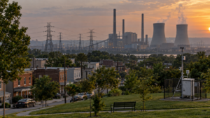 Illustrative image of a residential neighborhood, air-quality monitor and transmission lines near an unbranded fossil-fuel power plant at sunset.
