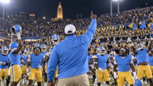Illustrative image of an anonymous football coach celebrating with players and a marching band in an unbranded blue-and-gold HBCU stadium.