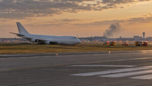 Illustrative image of an unbranded cargo aircraft beyond an airport runway as emergency vehicles respond, with smoke visible in the distance.